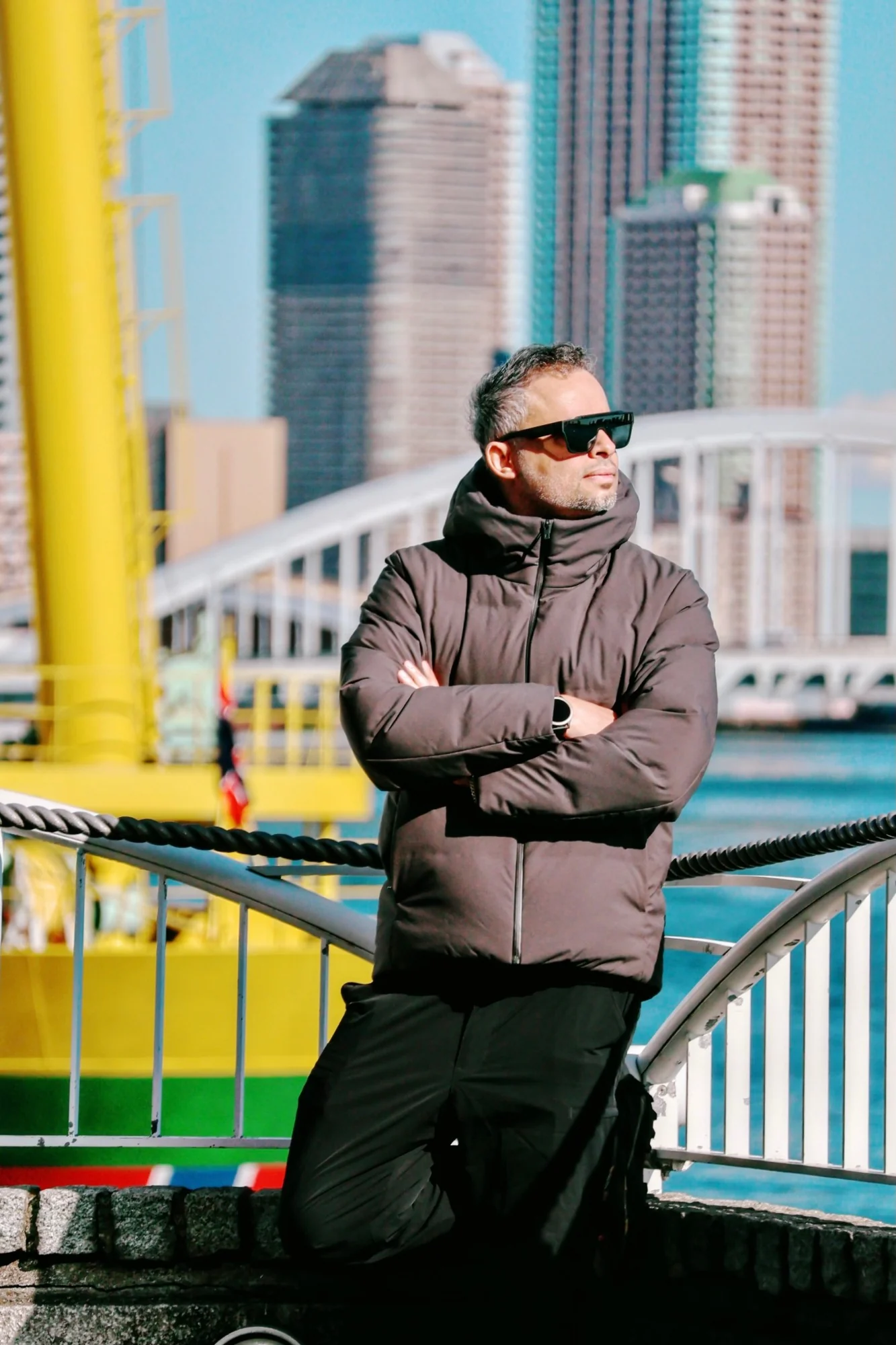 Alhasan Hosni in a dark puffer jacket and sunglasses, arms crossed, with a harbour and tall buildings behind him