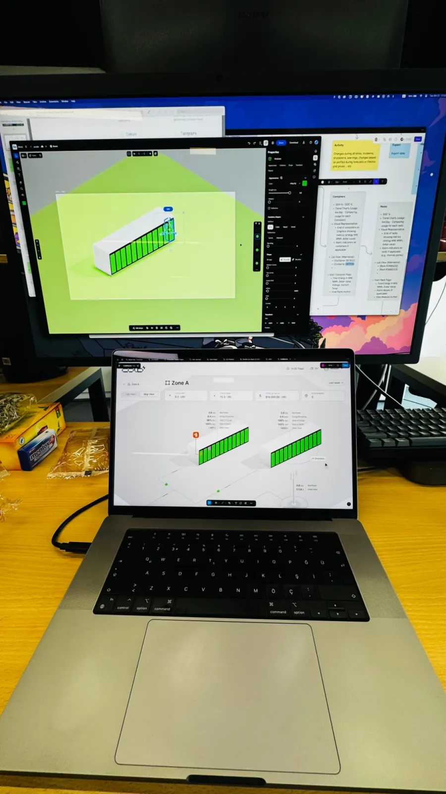 A big monitor with a 3D editor open on a green container model, above a laptop showing the Zone A map