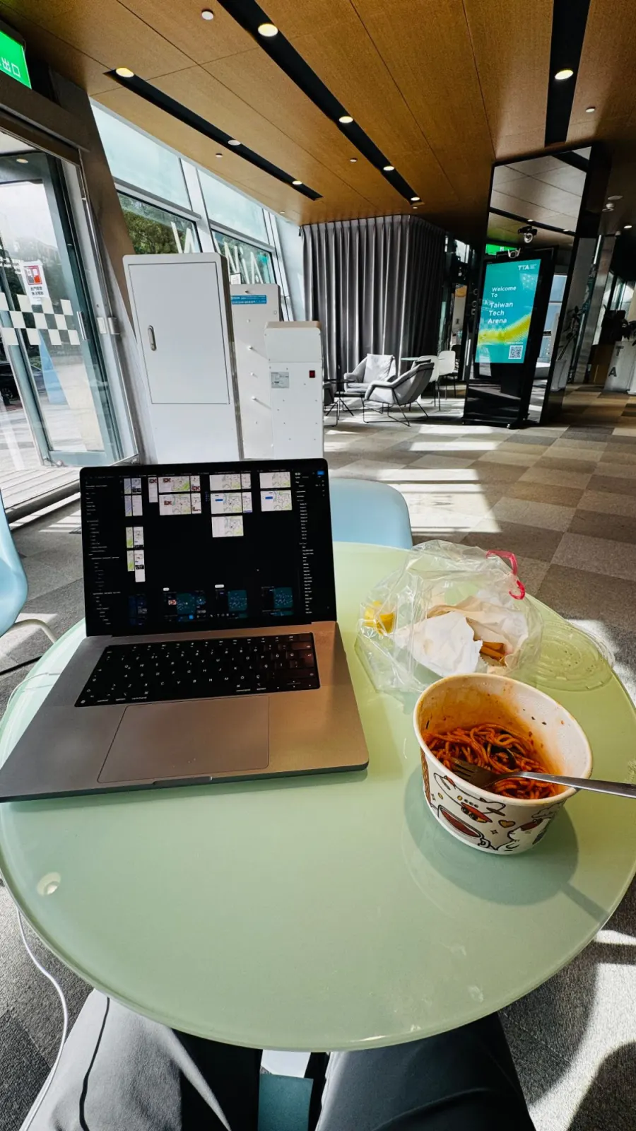 A laptop on a round table in a bright lobby, with design files open and a bowl of noodles beside it