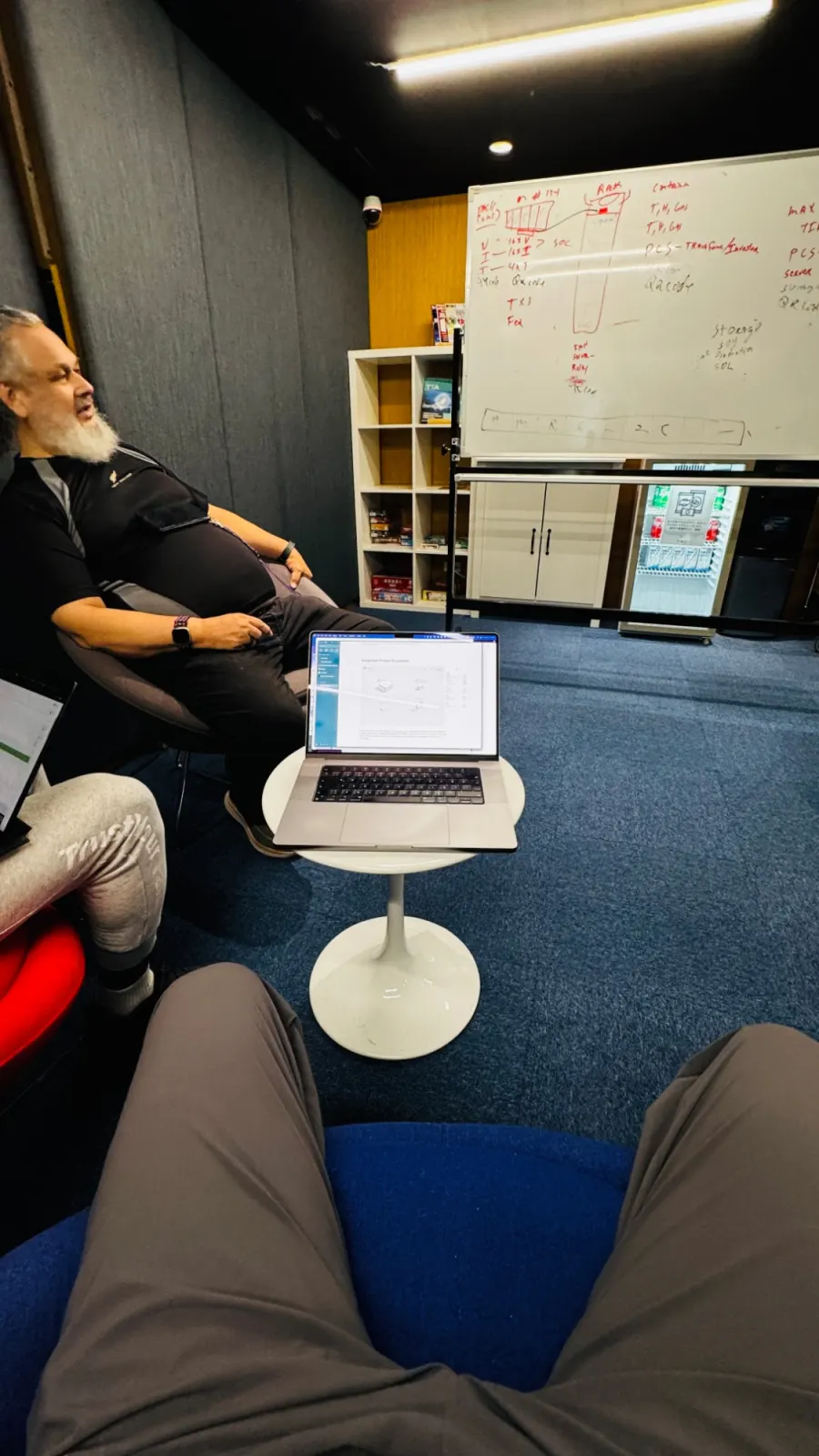 A meeting room with a whiteboard showing a rack, a PCS and state-of-charge notes, and a laptop on a small round table