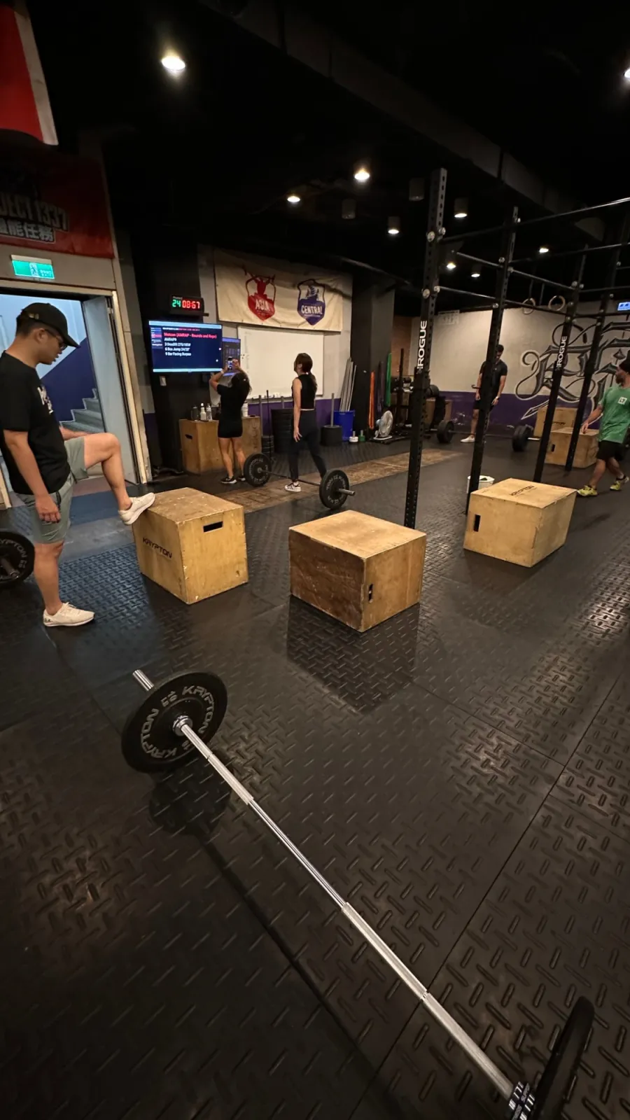 A CrossFit floor before class: plyo boxes, loaded barbells and a screen showing the workout