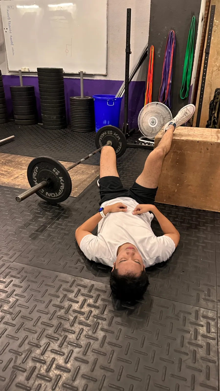 Someone lying on the floor of a gym with a leg up on a box, barbell beside them